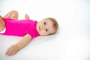 Wall Mural - cute, beautiful little girl in a pink bodysuit
 and a bandage lies on a white background, smiles and gnaws toys. beautiful baby. teeth are erupting. portrait of a baby. portrait of a beautiful girl