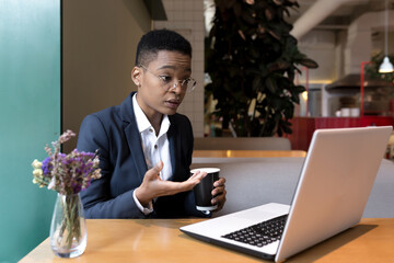 African-American business woman in a cafe makes a video call, thoughtful and focused talking to colleagues during lunch break, using a laptop and online communication