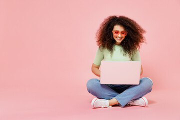 Poster - Full size body length fun young curly latin woman 20s wear casual clothes sit on floor in lotus pose hold use work on laptop pc computer isolated on plain pastel light pink background studio portrait