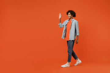 Wall Mural - Full body side view young smiling black student man 50s wearing blue shirt t-shirt waving hand walk go strolling isolated on plain orange color background studio portrait. People lifestyle concept