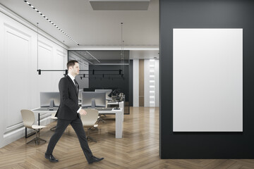 Poster - Thoughtful businessman walking in luxury coworking office interior with empty white mock up poster, furniture, wooden flooring, concrete walls, equipment and daylight. 