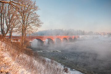 Wall Mural - Long red brick bridge over Venta river in misty, sunny, snowy winter morning with frosted trees, Kuldiga, Latvia