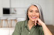 Close-up portrait on senior gray-haired woman in home interior, charming mature asian lady looking at camera and smiles, resting chin on hand, serene female spends weekend at home