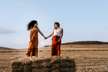 Young Women Looking At Each Other While Standing On Hay Bale In Farm
