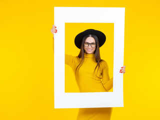 Happy woman holds a white frame in front of her face. Smiling girl. Girl in a yellow dress and a black hat. The lady poses as in the picture. A bright studio shot of a long-haired positive model.