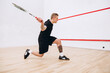 Full-length portrait of young sportive man in black uniform training, playing squash in sport studio