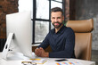 © Vadim Pastuh - Optimistic male office employee sitting at the desk in front of PC, looking at camera and smiling, successful young businessman in modern loft office, manager ceo in smart casual on the workplace