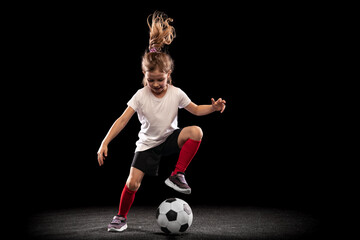 Wall Mural - Full-length portrait of playful girl, child in motion, action, training isolated over black background. Dribbling