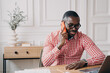 Young modern african businessman talking via mobile phone with client while working remotely