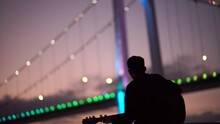 Silhouette Of Man Playing Guitar In Front Of A Bridge During Nightfall, Romantic Artist Singing In Outdoor
