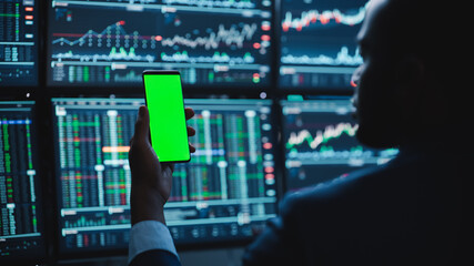 Poster - Financial Analyst Using Smartphone with Green Screen Chroma Key Mock Up Template and Working on Multi-Monitor Workstation with Stocks Charts. Businessman Works in Investment Bank in the Evening.