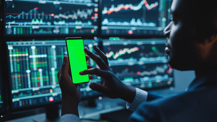 Poster - Financial Analyst Using Smartphone with Green Screen Chroma Key Mock Up Template and Working on Multi-Monitor Workstation with Stocks Charts. Businessman Works in Investment Bank in the Evening.