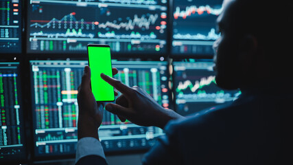 Poster - Financial Analyst Using Smartphone with Green Screen Chroma Key Mock Up Template and Working on Multi-Monitor Workstation with Stocks Charts. Businessman Works in Investment Bank in the Evening.