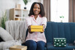 © sofiko14 - Attractive african woman sitting on couch with yellow gift box in hands and smiling on camera. Happy young lady enjoying festive mood and holidays tradition.