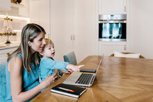 Boy Pointing At Laptop While Sitting On Mother's Lap Working In Kitchen