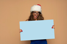 Christmas, X-mas, People, Advertisement, Sale Concept - Happy Woman In Blue Dress And Santa Helper Hat With Blank Blue Board On A Beige Background