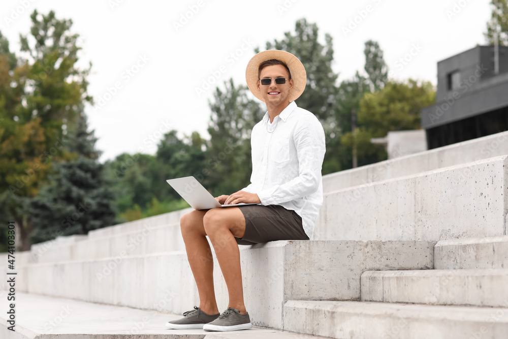 Handsome young man in sunglasses using laptop on stairs outdoors