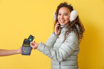 Canvas Print - Young woman in winter clothes making payment via banking terminal on color background