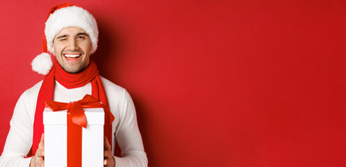 Concept of winter holidays, christmas and lifestyle. Handsome cheeky man in santa hat and scarf, holding present and smiling, winking at camera, standing over red background