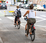 Fototapeta Miasto - Two lean and full-bodied women ride bicycles rented from a rental station along a dedicated cycle path in a modern city street