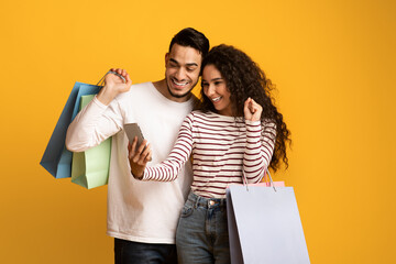 Wall Mural - Cashback. Joyful Arab Couple Holding Shopping Bags And Celebrating Success With Smartphone