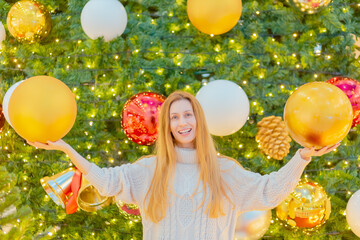 Wall Mural - A joyful lady is holding huge Christmas balls against the backdrop of an elegant sparkling Christmas tree. Festive poster. Congratulation