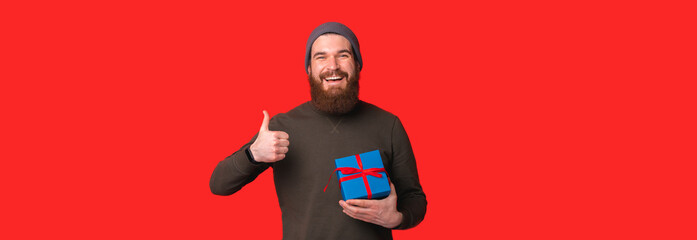 Wall Mural - Banner size photo of young cheerful man with beard holding blue gift box and showing thumb up.