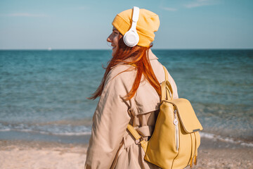 Lifestyle portrait of stylish Caucasian red hair woman wearing headphones listening music or podcast from smartphone application against the sea