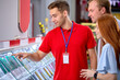 © alfa27 - Nice Caucasian Male Consultant In Red Uniform Showing Representing New Phone In Store Tech Shop To Customer, Showing Through Glass, Talk About Advantages And Characteristics Of Mobile Phone
