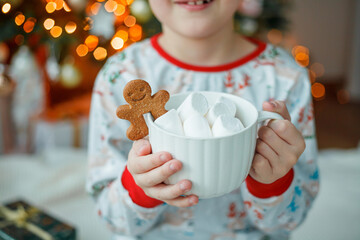 Adorable 7 year old boy in pajamas drinks hot cocoa with marshmallows in white mug with gingerbread man cookies near the Christmas tree. New Year's lights. Winter holidays.