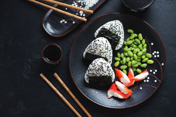 Wall Mural - Black plate with japanese onigiri, crab meat and edamame, top view on a black stone background, horizontal shot