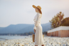 Stunning Blonde Woman In Summer Beach Outfit Relaxing Outdoors Against Sea Resort And Palm Trees On The Background. A Fashionable Romantic Young Adult Lady Wearing A Trendy Vintage Straw Hat, White
