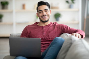 Wall Mural - Smiling arab guy reclining on sofa with laptop