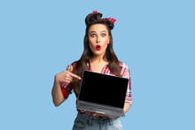 Shocked Pinup Woman In Retro Outfit Pointing At Laptop With Empty Screen On Blue Background