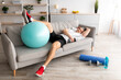 Fatigue mature man resting on sofa after active hard training. Tired sportsman with dumbbells and fitness ball on couch