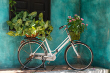 White Vintage Bike With Basket Full Of Flowers Next To An Old Building In Danang, Vietnam, Close Up