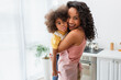Happy african american mother in apron hugging child sticking out tongue in kitchen.