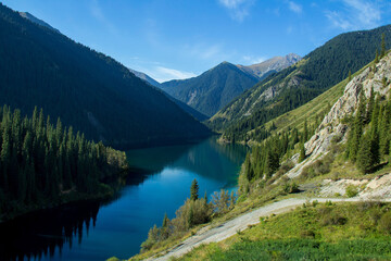 Wall Mural - In a beautiful alpine gorge there is a large lake Kolsai, the lake is surrounded by mountain ranges with peaks, a spruce forest grows on the slopes, a road in the foreground, summer, sunny