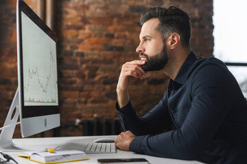 Wall Mural - Focused and concerned man in smart casual shirt looking at laptop screen. Serious caucasian guy sitting at the desk and feeling doubts, while solving difficult tasks