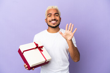 Poster - Young Colombian man holding a gift isolated on purple background counting five with fingers