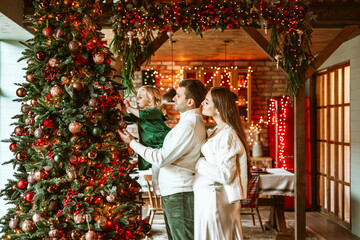 family of four parents pregnant mom and dad and two siblings little boy and girl in stylish clothes in chalet are ready to welcome Christmas and New Year near Christmas tree with gift box presents