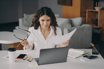 Wall Mural - Smiling glad italian woman paying bills online at home while sitting on living room