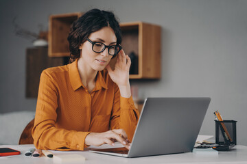 Wall Mural - ocused Spanish woman in glasses sits at desk and typing on laptop during remote work at home