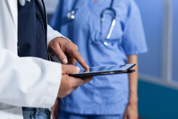 Close up of doctor pointing at tablet display for medical practice, working late. Medic using modern device with touch screen and talking to nurse about healthcare system overtime.