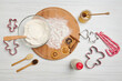 Flat lay of ingredients for preparing dough and gingerbread cookies, cutting molds, bottle with plant based milk, candy canes, glass jar with honey and a bowl with flour on a round wooden board