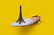 A right hand in a white medical glove holds a miniature metal Eiffel Tower.Torn hole in yellow paper.The concept of hygiene,self-isolation during quarantine measures to combat the covid-19 coronavirus