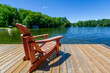 A brown Adirondack chair on a wooden pier facing a lake in Muskoka, Ontario Canada. Across the calm water are few cottages nestled among green trees.