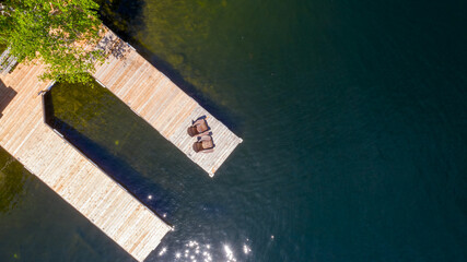 Wall Mural - Aerial view of a cottage wooden pier on a lake in Muskoka, Ontario Canada. Two brown Adirondack chairs are visible on the dock facing the blue waters of the lake.