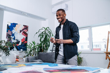 Carefree perky black man standing next to a big canvas with painting on a table