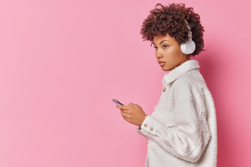 Wall Mural - Sideways shot of thoughtful curly haired young woman listens music via headphones holds mobile phone uses modern gadget dressed in white fur coat isolated over pink background blank copy space